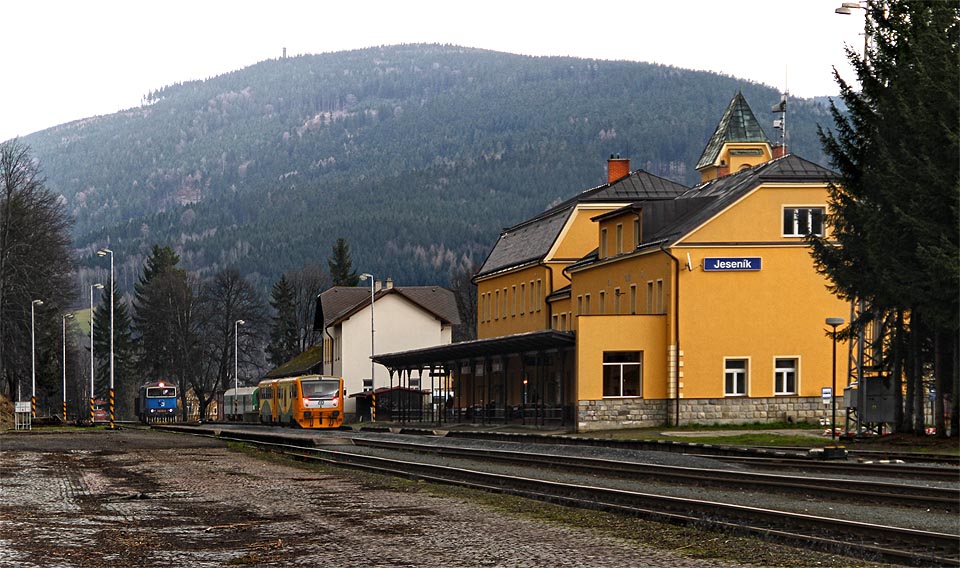 Stanice Jeseník se spěšným (814) a manipulačním (753) vlakem přišedšími v těsném sledu za sebou z Mikulovic. V pozadí majestátní kopec Zlatý chlum.