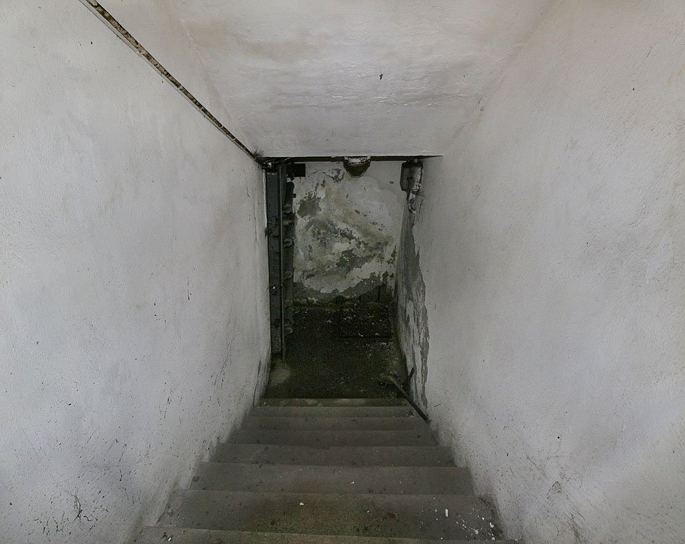 Vstup ze západní strany není o nic malebnější. K podzemním pancéřovým dveřím vede jen několik zvlhlých schodů.